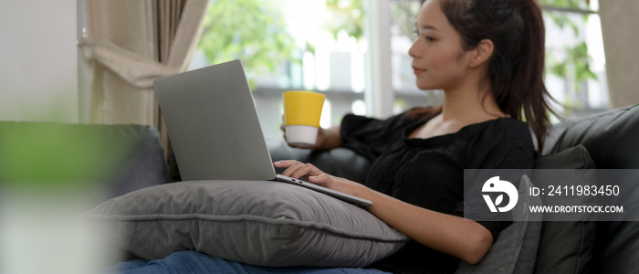 Young female lying relaxed on sofa and holding a cup of coffee while using laptop