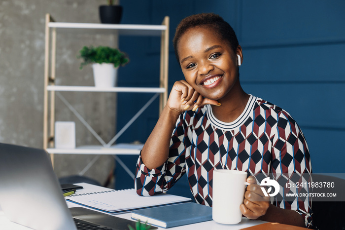 Portrait of an African woman posing at the office desk and looking at the camera while holding a cup