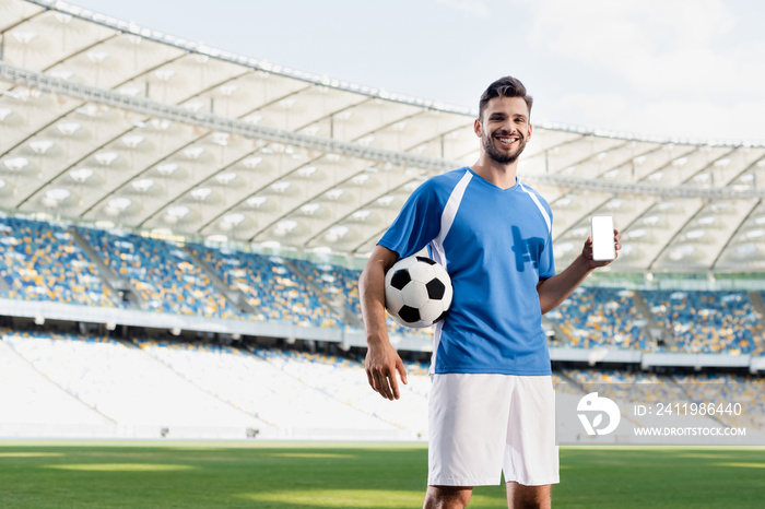 smiling professional soccer player in blue and white uniform with ball showing smartphone with blank