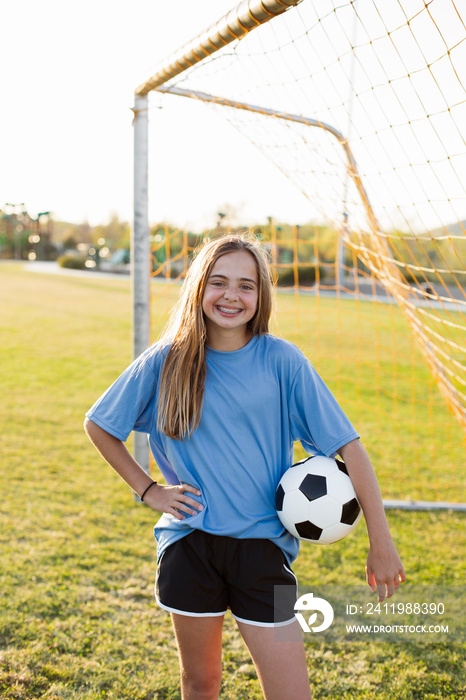 Portrait of smiling player with soccer ball standing on grassy field against clear sky