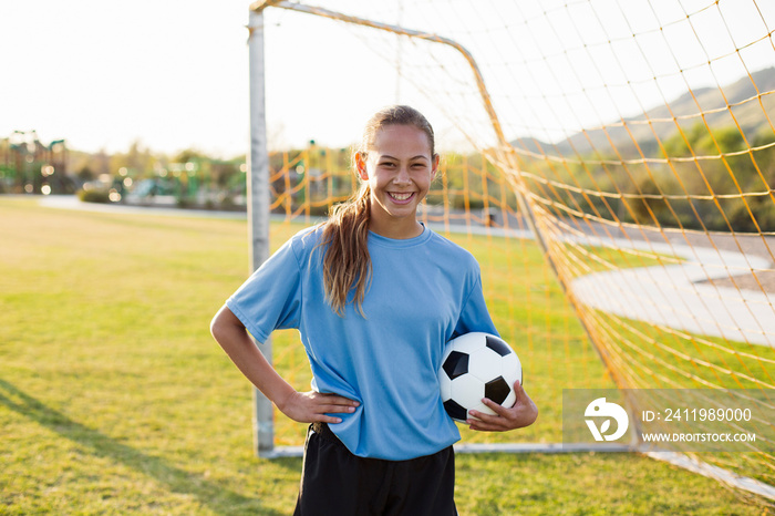 Portrait of smiling player holding soccer ball while standing on grassy field against clear sky