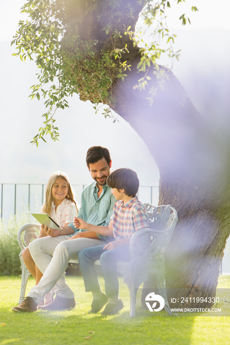 Family using digital tablet on bench below sunny tree in backyard