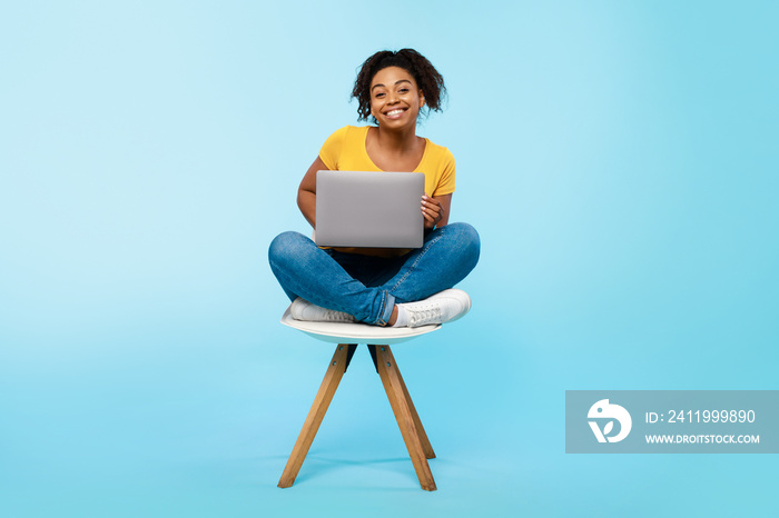 Cheery young black woman working online, sitting on chair and using laptop on blue studio background