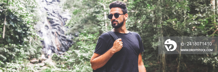 Handsome young stylish man in black t-shirt and sunglasses is engaged in trekking in the green jungl