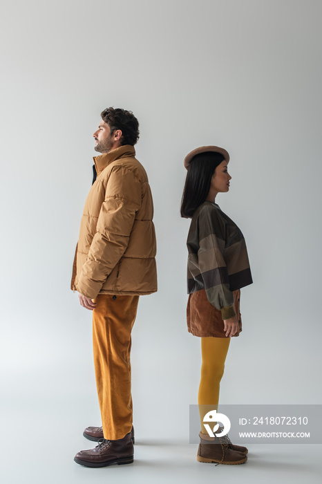 full length view of interracial couple in beige and yellow clothes standing back to back on grey