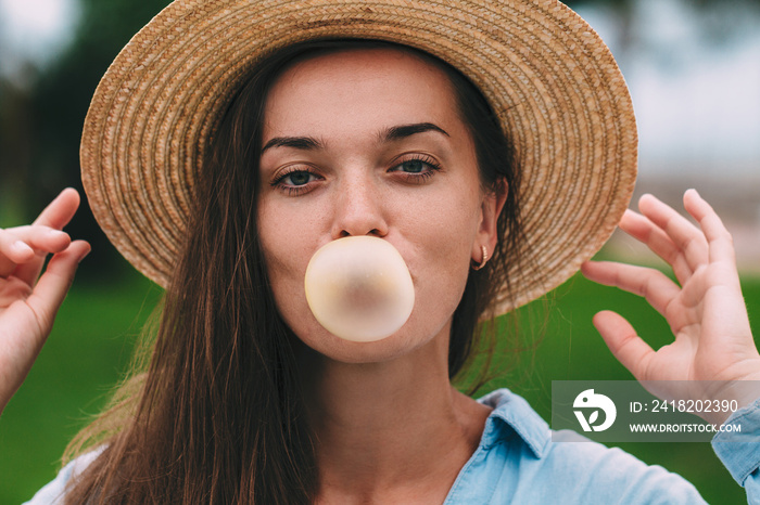 Young cheerful happy hipster woman in hat blowing bubble of chewing gum outdoor