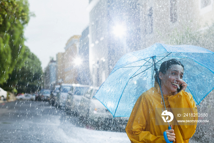 Happy woman with umbrella talking on smart phone on rainy street