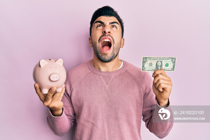 Young hispanic man holding one dollar banknote and piggy bank angry and mad screaming frustrated and