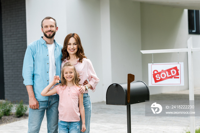 happy man standing with wife and daughter near house and board with sold letters