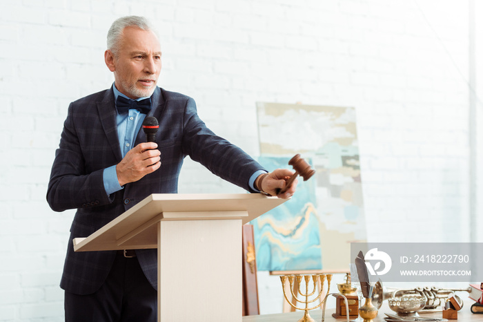 handsome auctioneer in suit talking with microphone and holding gavel during auction