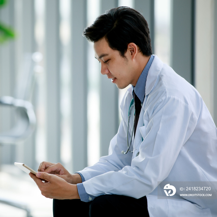 Handsome young Asian doctor in clinic hygiene gown smile and happy to sit for relaxing and messaging