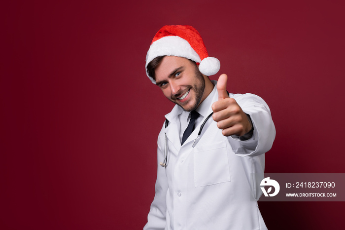 Young handsome doctor in white uniforme and Santa Claus hat standing in studio on red background smi