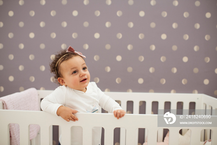 Cute baby girl looking away while standing in crib against wall at home