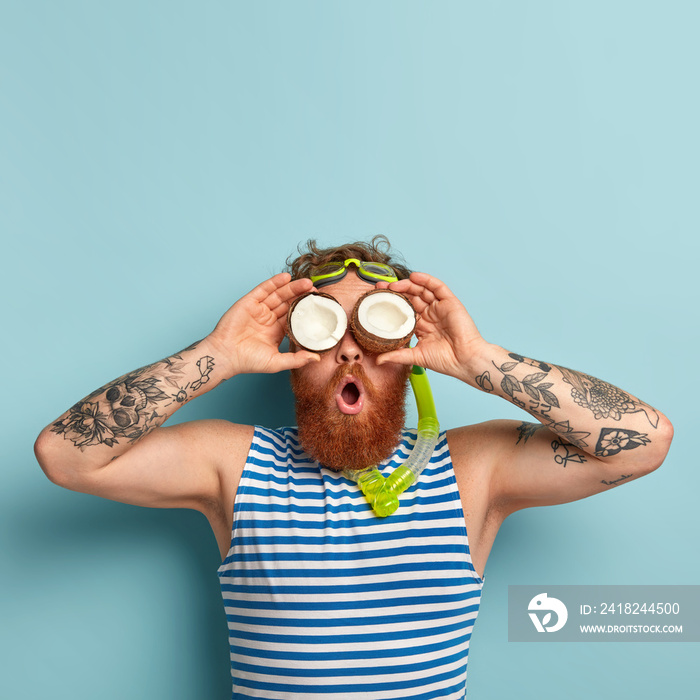 Photo of funny surprised guy covers eyes with coconuts, has thick ginger beard, uses snorkel mask fo