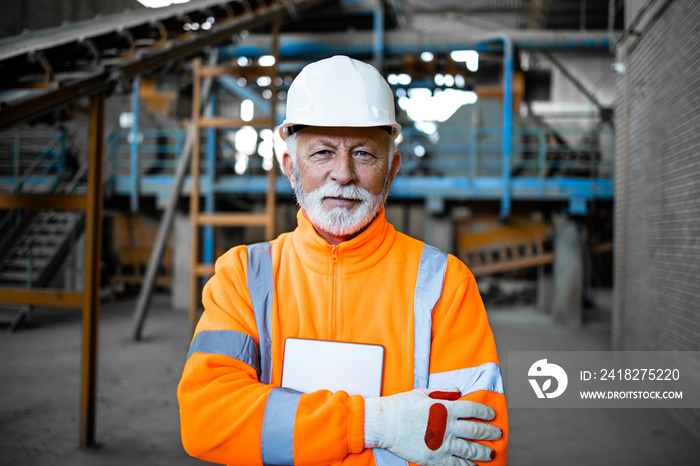 Portrait of an experienced senior industrial worker in safety equipment standing in factory interior