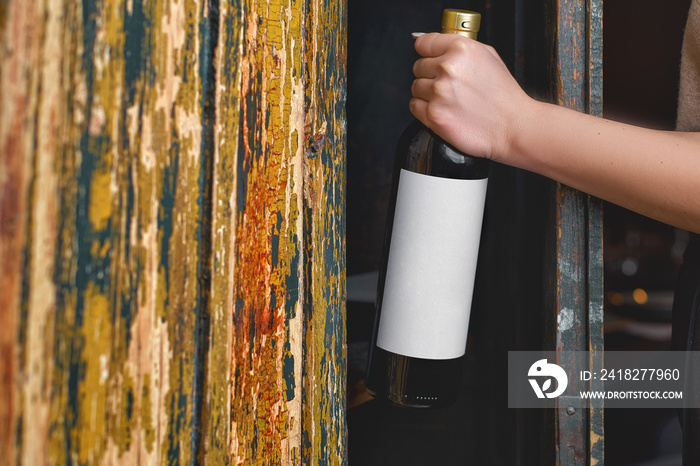 女性手拿着带有白色空白标签的红葡萄酒瓶，靠着旧葡萄酒架的特写