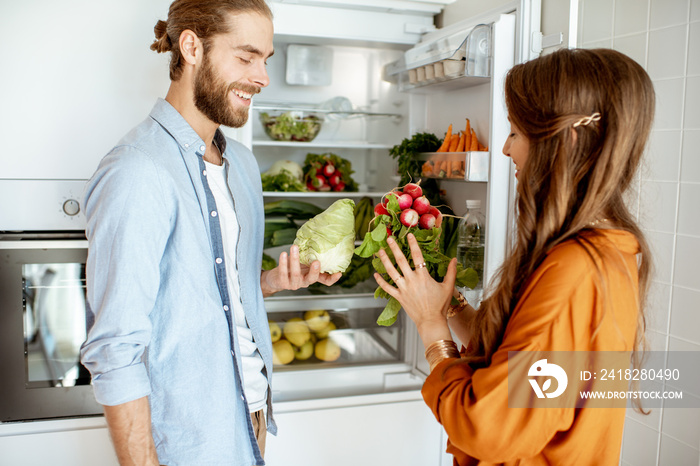 Young vegan couple choosing what to cook, taking fresh vegetables from the refrigerator at home