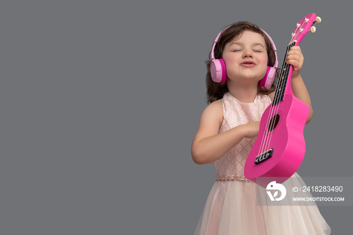一个美丽有趣的小女孩在工作室里用灰色的背景弹着粉红色的玩具吉他。
