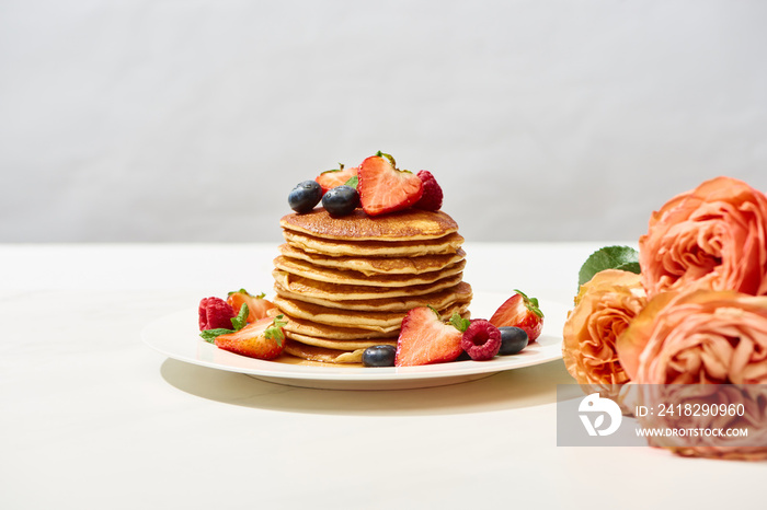 selective focus of delicious pancakes with blueberries and strawberries on plate near rose flowers o