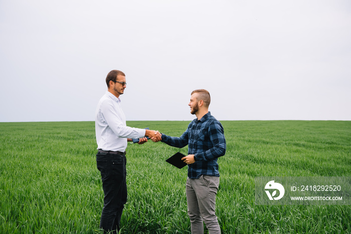 Two farmer standing in a green wheat field and shake hands.