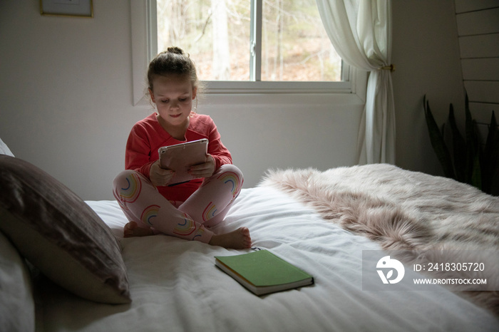 Girl using tablet computer while sitting on bed against window at home