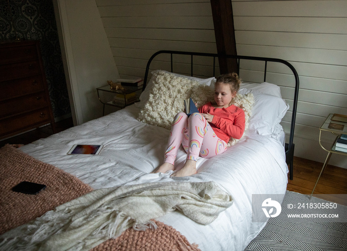 High angle view of girl reading book while relaxing on bed against wall at home