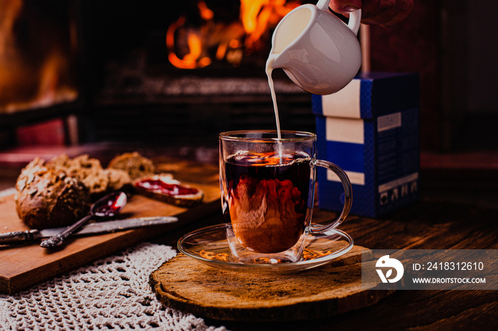 Pouring milk from porcelain milk jug into cup filled with tea - white tablecloth and fire background