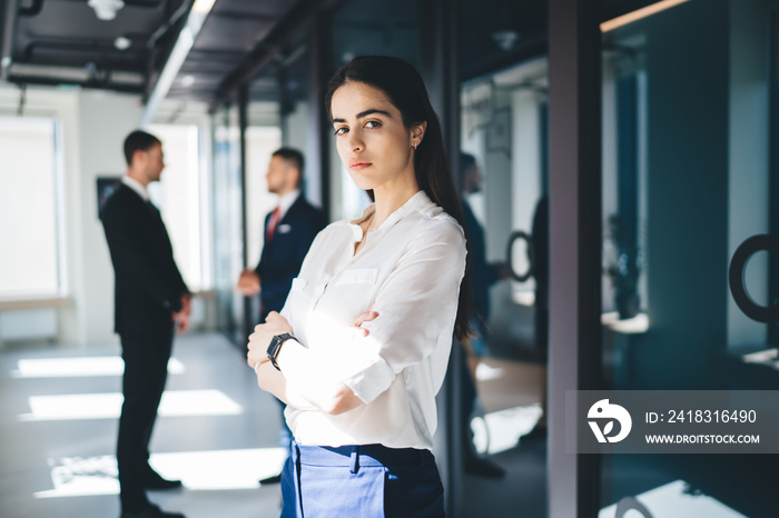 Serious young ethnic businesswoman standing in office corridor with crossed arms