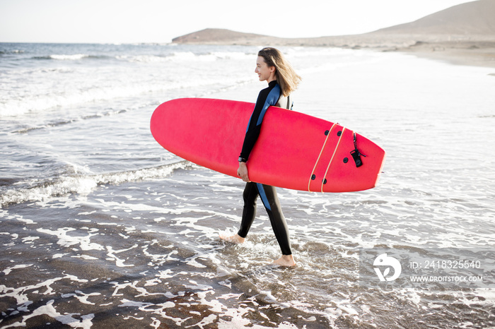 Young woman in wetsuit walking with surfboard on the beautiful wild beach on a sunset. Water sport a