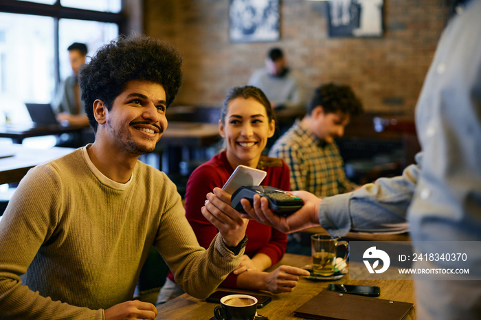 Young man making contactless payment with smart phone while being with his girlfriend in a cafe.