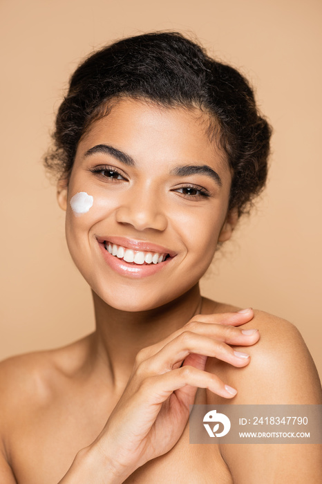 joyful african american woman with face cream on cheek isolated on beige