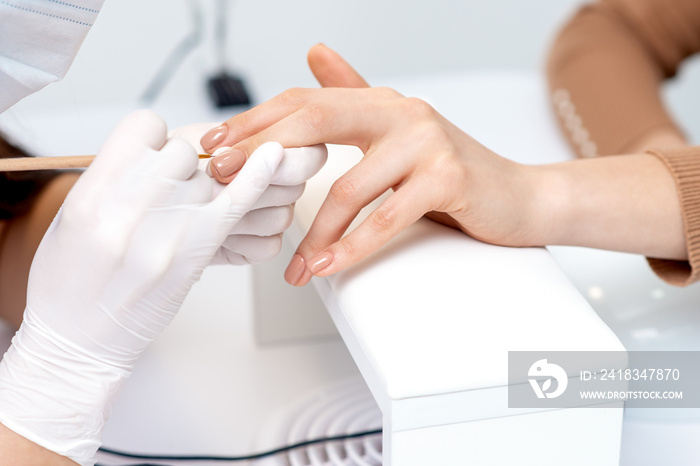 Manicure master in protective gloves applying beige nail polish on female nails in beauty salon