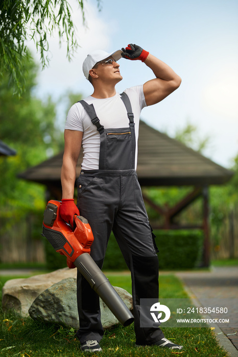 Full length portrait of handsome caucasian man in overalls, cap and gloves standing at garden with l