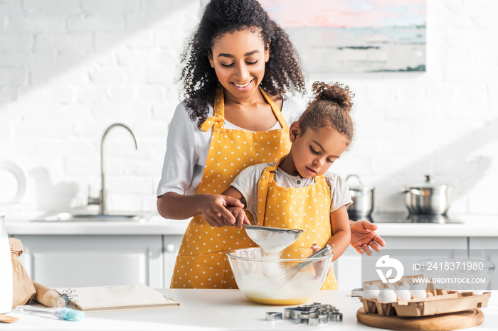 smiling african american mother and daughter preparing dough and sieving flour in kitchen