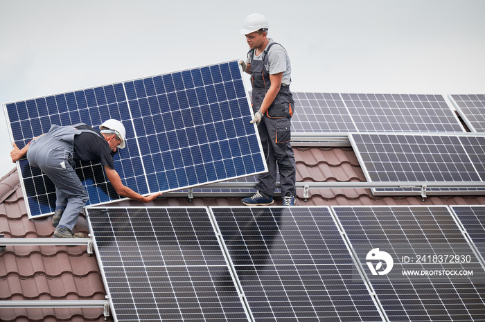 Men technicians carrying photovoltaic solar moduls on roof of house. Engineers in helmets installing