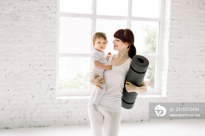 运动型漂亮年轻母亲，手拿瑜伽垫，带着穿着白色运动服的小女婴，站着