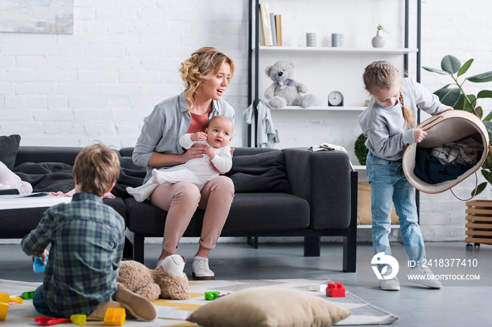 exhausted mother with infant child sitting on couch and looking at naughty siblings playing at home