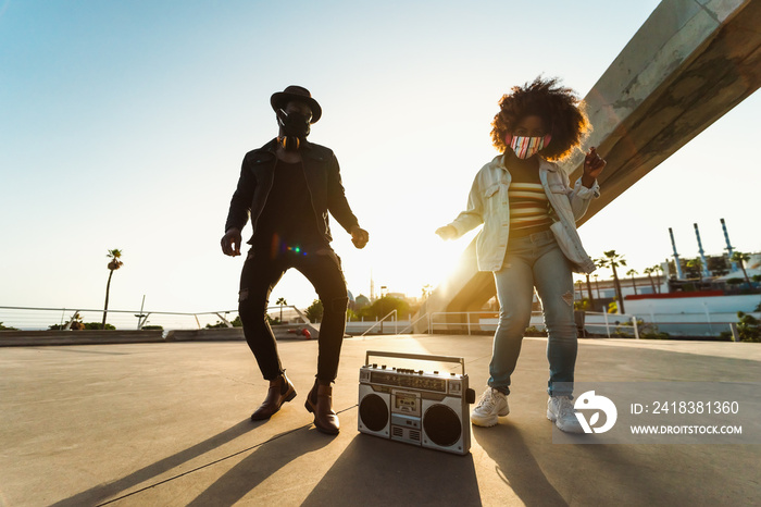 Young Afro friends wearing face mask dancing outdoor while listening to music with wireless headphon