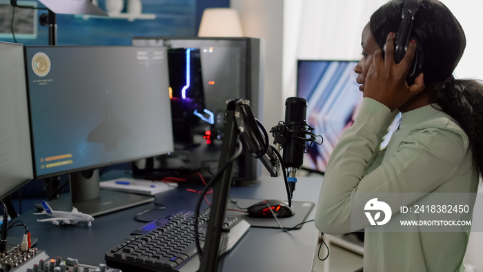 African woman streamer cyber putting on headphones performing space shooter video game on RGB powerf