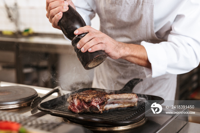 Image of adult man chief in white uniform cooking meal with meat and vegetables, at kitchen in resta