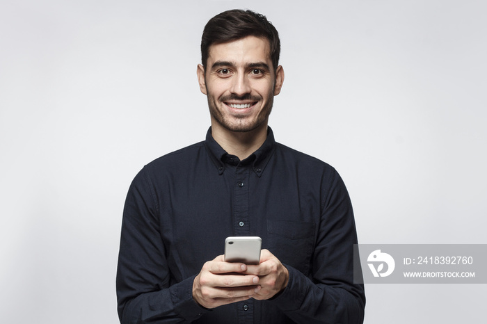 Young business man standing isolated on grey background, looking at camera, holding phone with both 