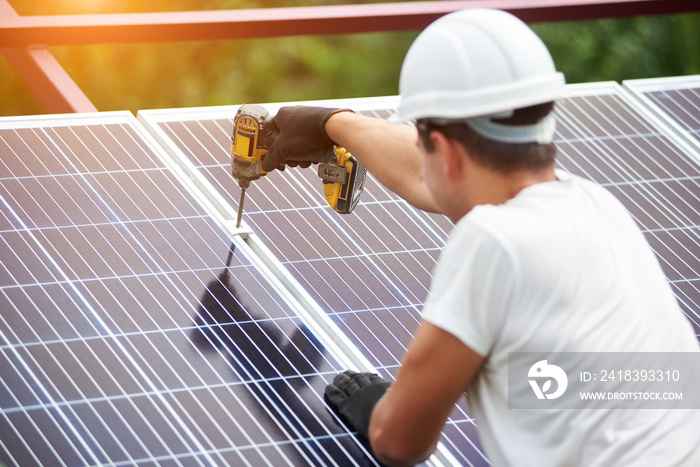 Back view of young technician in helmet connecting solar photo voltaic panel to metal platform using