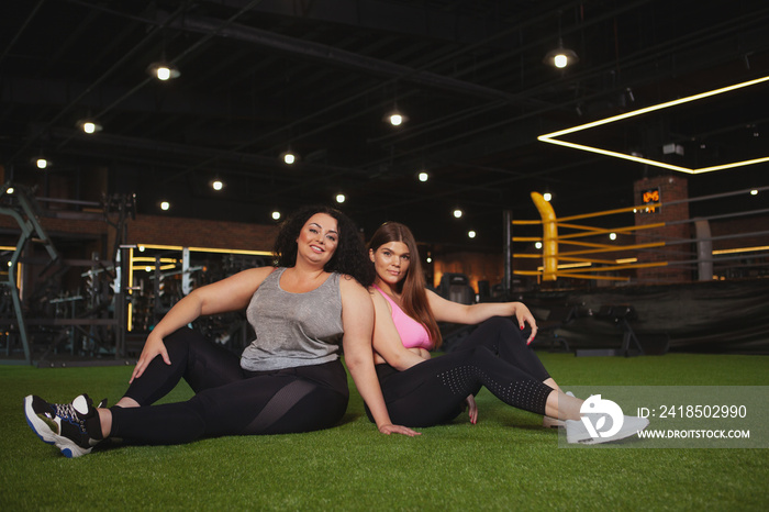 Two beautiful healthy happy plus size fitness women resting at the gym after exercising, copy space.