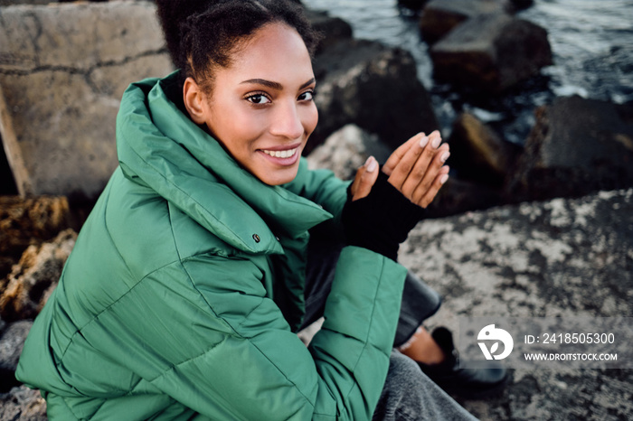 Pretty African American girl in down jacket warming hands happily looking in camera on stones by the