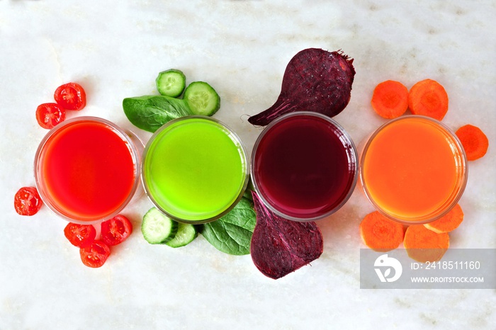 Four types of juice with scattered vegetables, above view over a white marble background