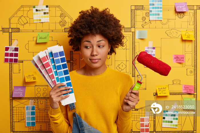 Thoughtful indecisive woman looks at color samples, holds paint roller, thinks about refurbishment o