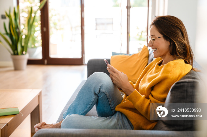 Beautiful woman smiling and using cellphone on sofa in apartment