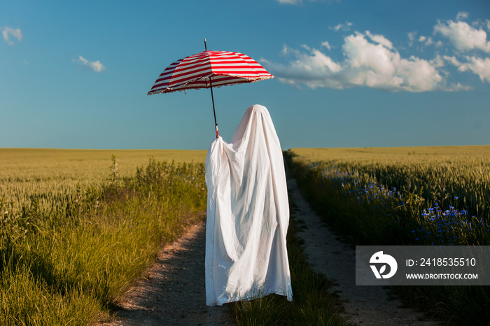 cute ghost in a bed sheet with umbrella on countryside road near a wheat field