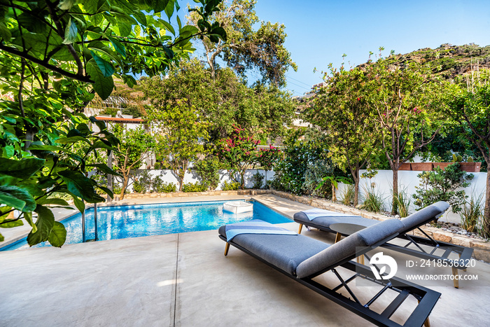 Pool area of a house or villa, with private swimming pool and two sun beds, greenery around. 