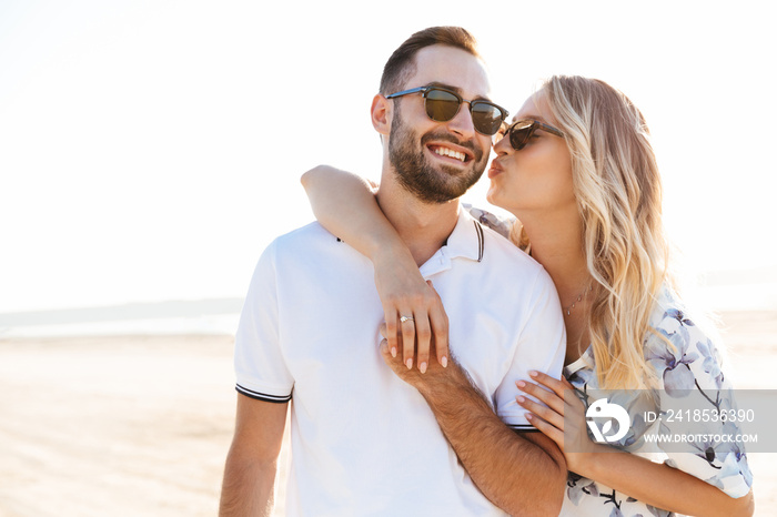 Photo of blonde young woman kissing and hugging handsome man while walking on sunny beach